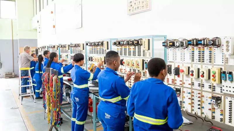 Apprentices in blue overalls with reflective stripes work along a wall of electrical control panels with switches, relays and coloured wires in a South African training centre