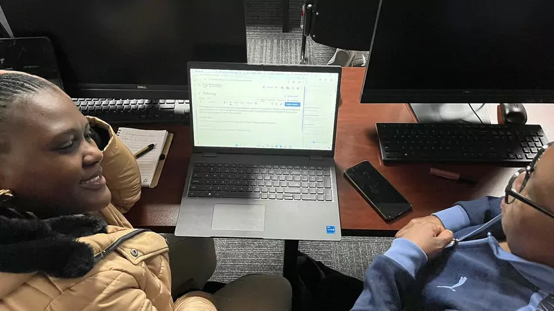 Two people seen from above sit at a desk in a university computer lab, a woman in a tan puffer jacket smiling at the centre laptop, with desktop monitors, a keyboard, a notebook and a phone on the desk