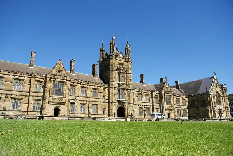 The sandstone Main Quadrangle building of the University of Sydney with its clock tower, across a wide green lawn under a blue sky