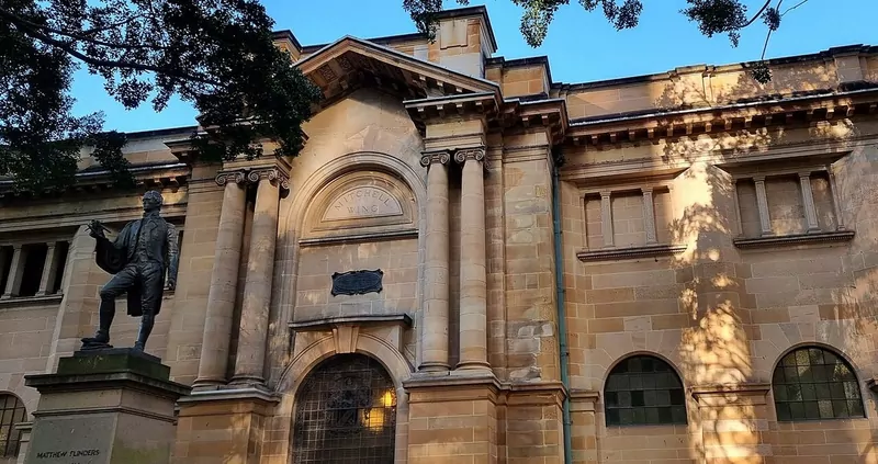 The sandstone Mitchell wing of the State Library of New South Wales with tall columns, an arched doorway and a bronze statue of Matthew Flinders on the left
