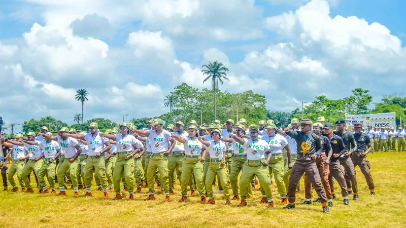 Rows of corps members in white NYSC T-shirts, khaki trousers and caps raise their fists on a grass field with palm trees and a blue sky behind them