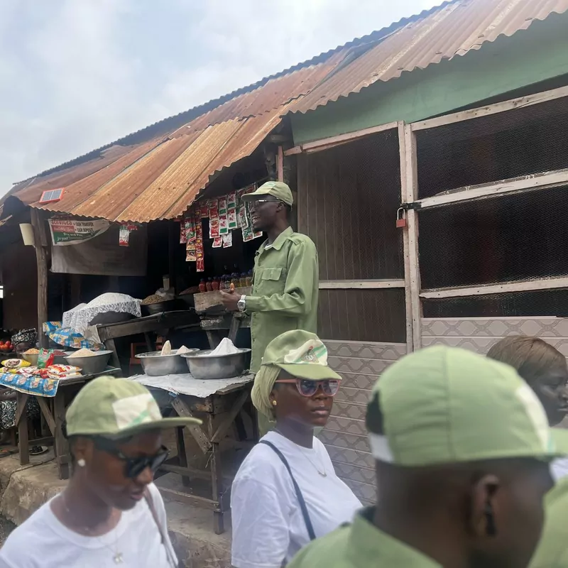 A corps member in a green khaki uniform and cap stands at a market stall with a rusty tin roof, with other corps members in caps and sunglasses in the foreground
