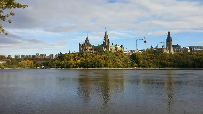 Parliament Hill in Ottawa seen across the Ottawa River, with green and autumn-coloured trees below the buildings and clouds above
