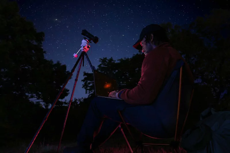 A man in a cap sitting outdoors at night with a MacBook Pro on his lap next to a telescope under a starry sky