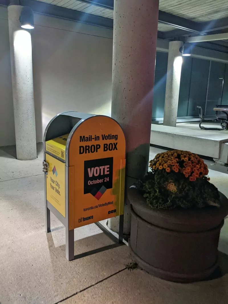 A yellow metal Mail-in Voting Drop Box from the City of Toronto, with a Vote sign, standing outside a community centre beside a pot of flowers