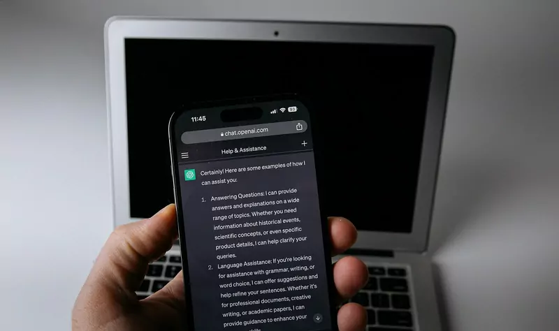 A hand holds a smartphone showing a ChatGPT conversation in front of an open laptop