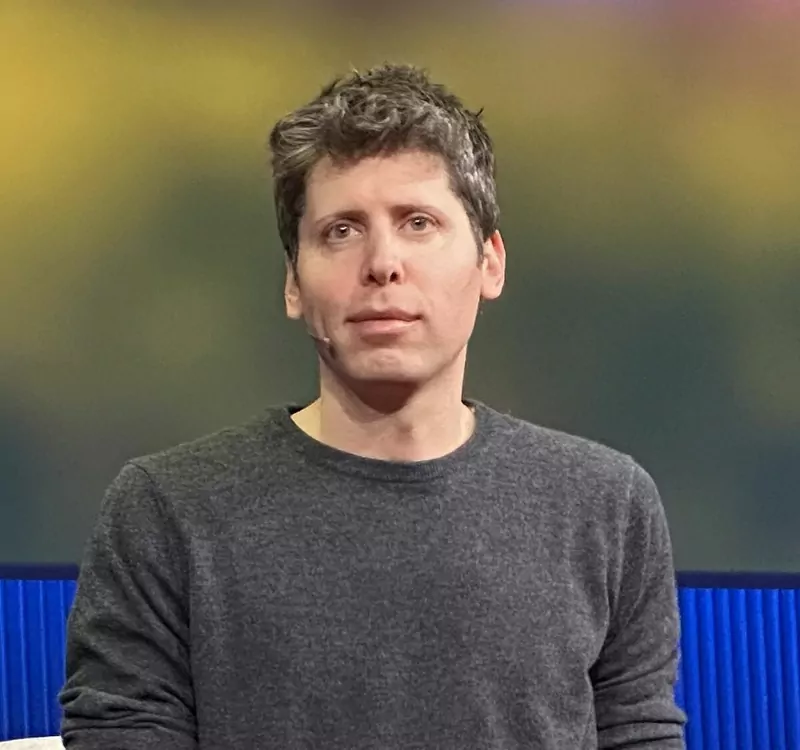 Sam Altman seated on a stage in a gray sweater and jeans