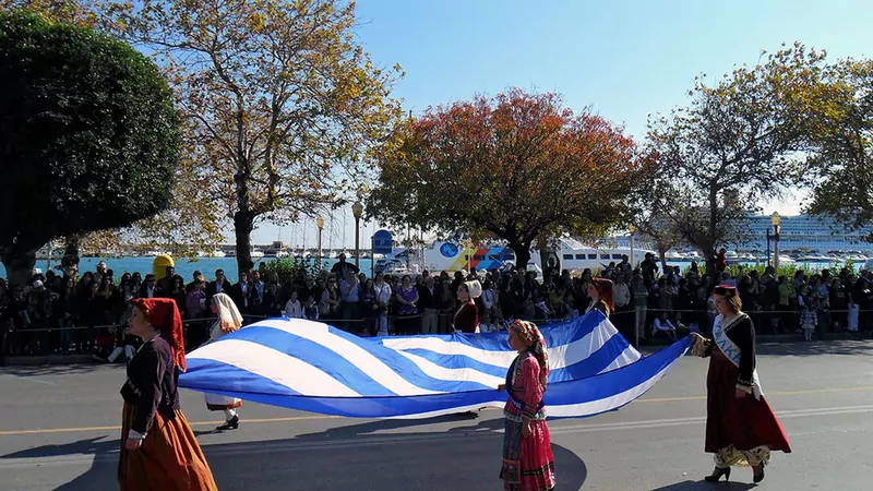 Women in traditional Greek costumes with long skirts carry a huge blue and white Greek flag along a seafront street lined with spectators in Rhodes