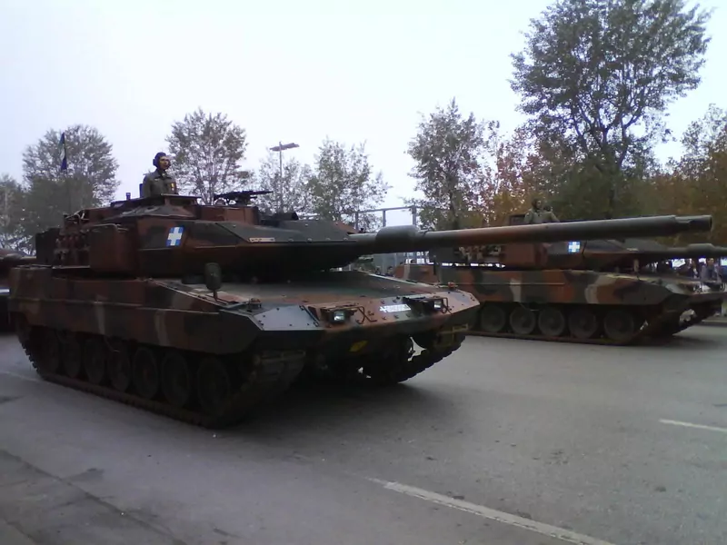 Camouflaged Leopard 2 tanks with soldiers in the turrets drive along a wide avenue during a parade, with autumn trees behind