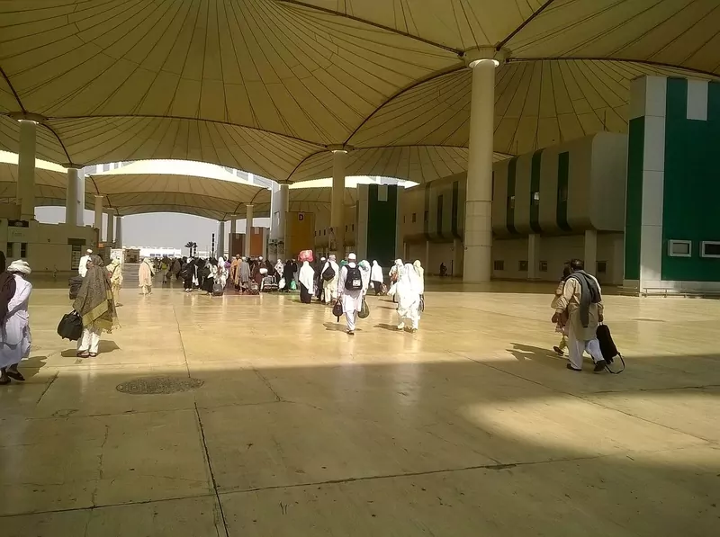 Travelers in white and light clothing walking with bags across a wide plaza under a large tent-style roof at an airport terminal