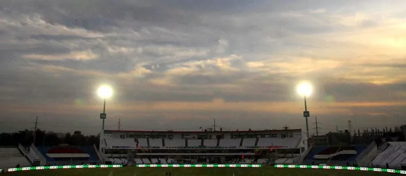 Rawalpindi Cricket Stadium at dusk with two floodlights lit and an almost empty stand under a cloudy sky