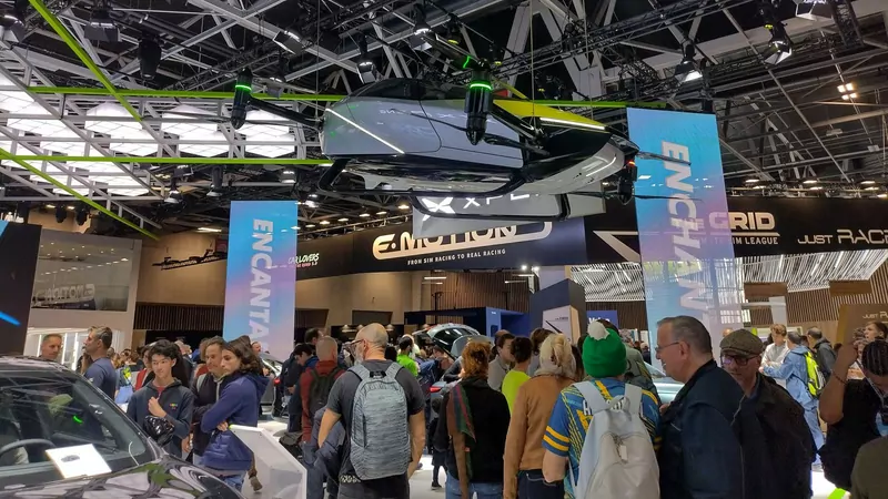 Crowds walk under a futuristic flying car prototype hanging from the ceiling of an exhibition hall full of car stands