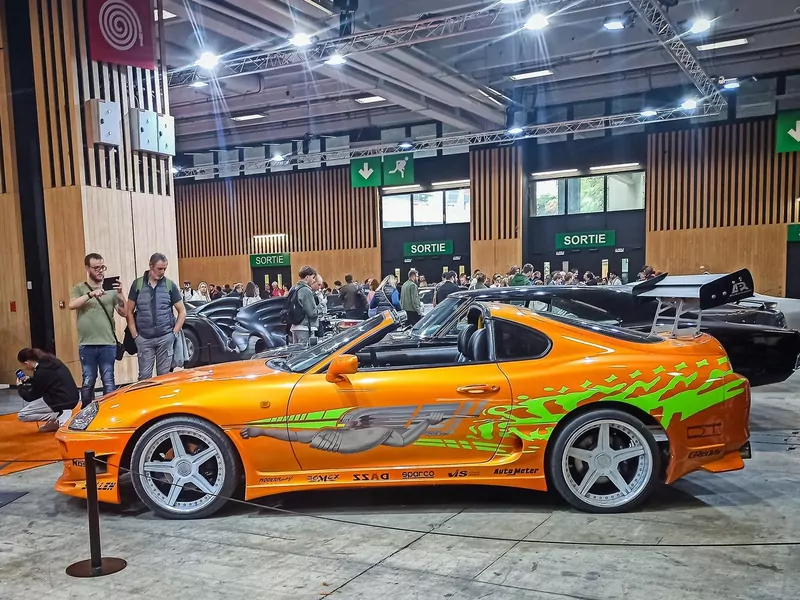 An orange sports car with green graphics on display in an exhibition hall, with visitors taking photos of other film cars behind it