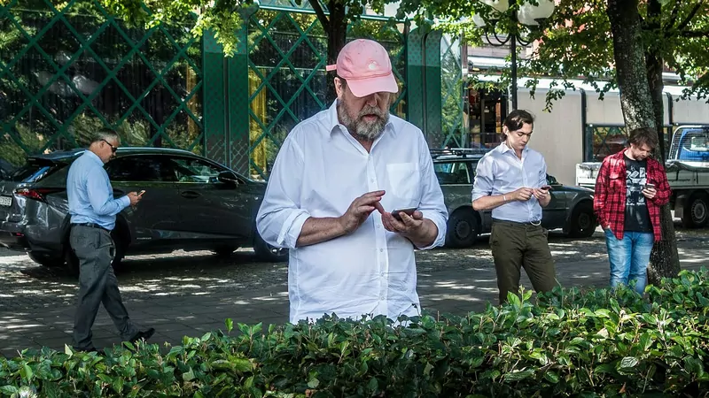 A man in a pink cap and white shirt taps his phone behind a hedge while three other men on the pavement also play on their phones