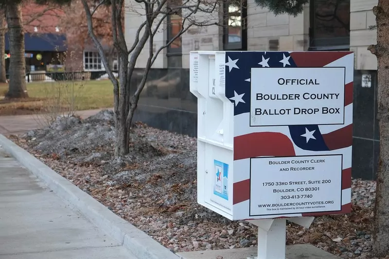 A white official ballot drop box with a stars and stripes design and the Boulder County address on its side, on a sidewalk