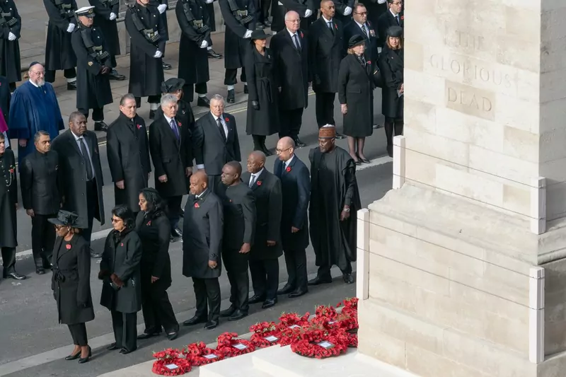 Dignitaries in dark coats standing in rows beside the Cenotaph in Whitehall, with red poppy wreaths laid at its base