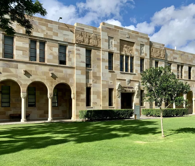 A two-storey sandstone university building with carved panels, arched cloisters on the left and a green lawn in front