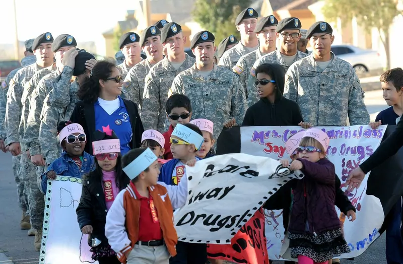 Young pupils in paper headbands reading No drugs carry hand-painted banners in a parade, with soldiers walking behind them