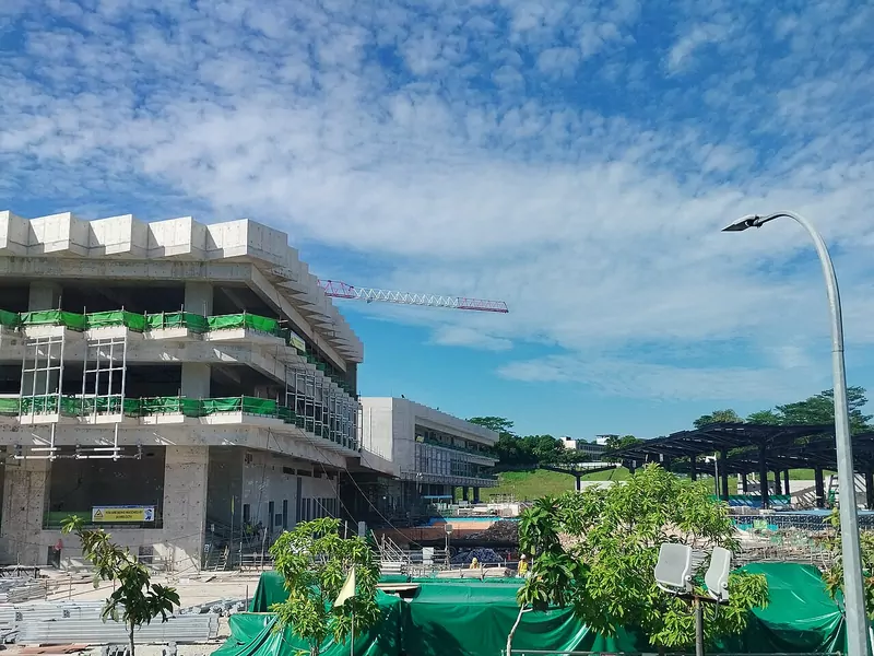 A concrete station building under construction with green safety netting, a tower crane and a street lamp against a blue sky with thin clouds, with green sheeting and trees in the foreground