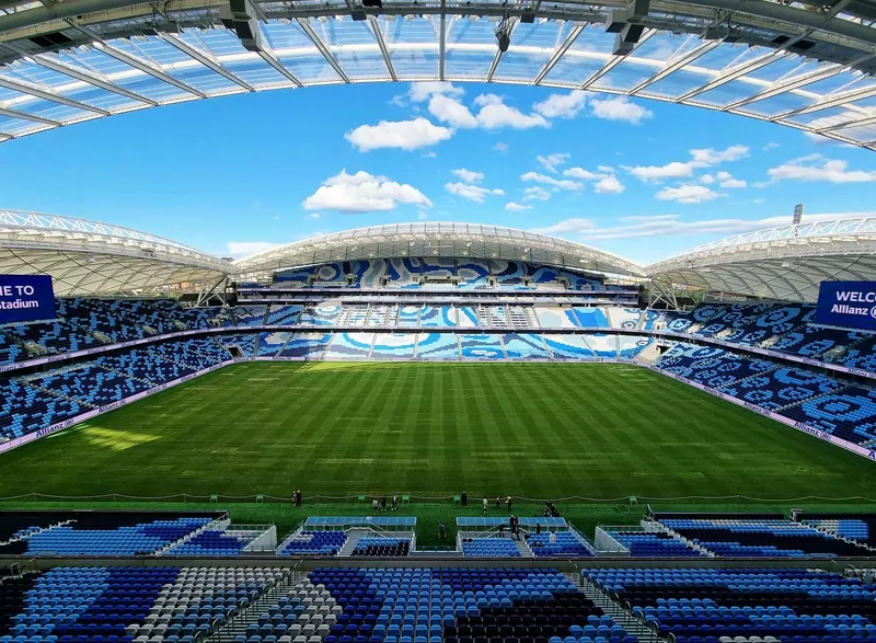 The inside of Sydney's Allianz Stadium on a sunny day, with blue and white seats in curved stands, a glass-panelled roof and a green pitch with mowing stripes