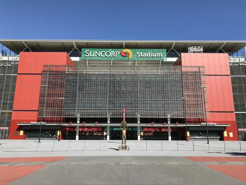 The red and orange front of Suncorp Stadium in Brisbane under a clear blue sky, with a green sign above the entrance and a bronze statue in the plaza