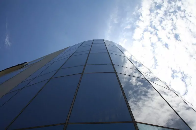Looking up at a curved glass tower of a South African Revenue Service office building, its dark panes reflecting white clouds against a deep blue sky
