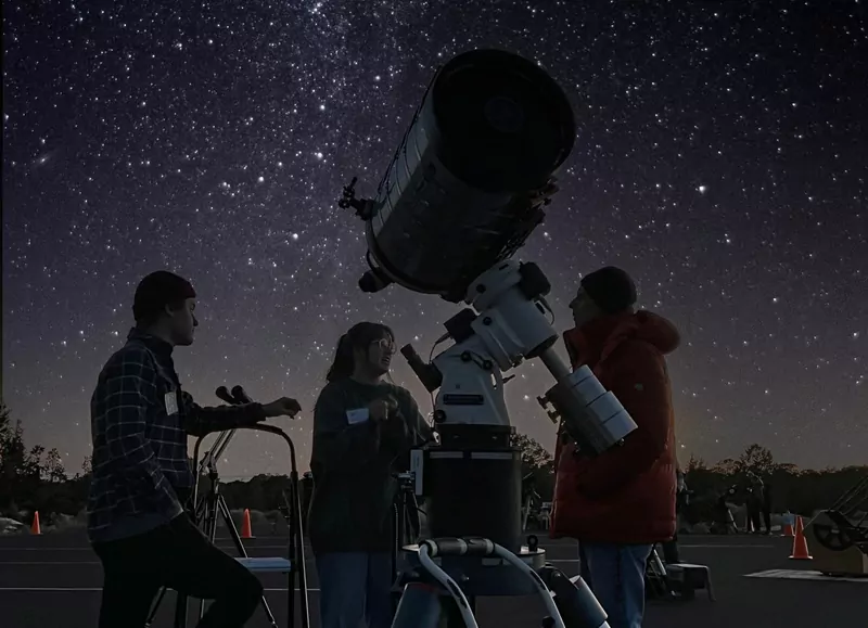 Three people standing around a large telescope under a sky full of stars