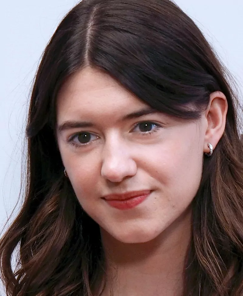 A close-up portrait of a young woman with long dark hair parted in the middle and small silver hoop earrings, in front of a pale background