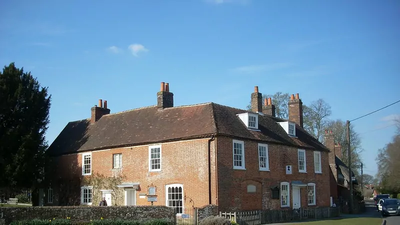 A two-storey red-brick house with a tiled roof and tall chimneys beside a village road, under a blue sky