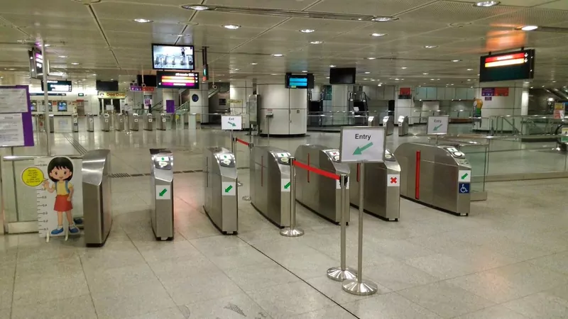A row of silver fare gates with green entry arrows in a quiet Dhoby Ghaut MRT station concourse, with a cardboard cut-out of a girl beside a height chart at the left