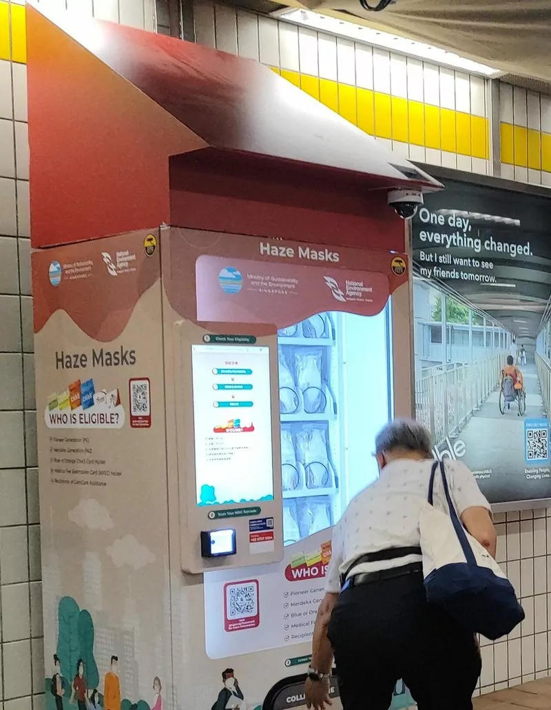 A tall red and cream vending machine labelled Haze Masks, with the Ministry of Sustainability and the Environment and National Environment Agency logos, a touchscreen, an eligibility list and an older man in a white shirt bending to collect a pack from the dispenser