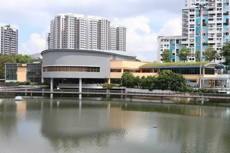 A community club building beside a calm pond in Bukit Panjang, with a curved grey upper storey, orange walls and a green roof terrace, and tall flats rising behind it
