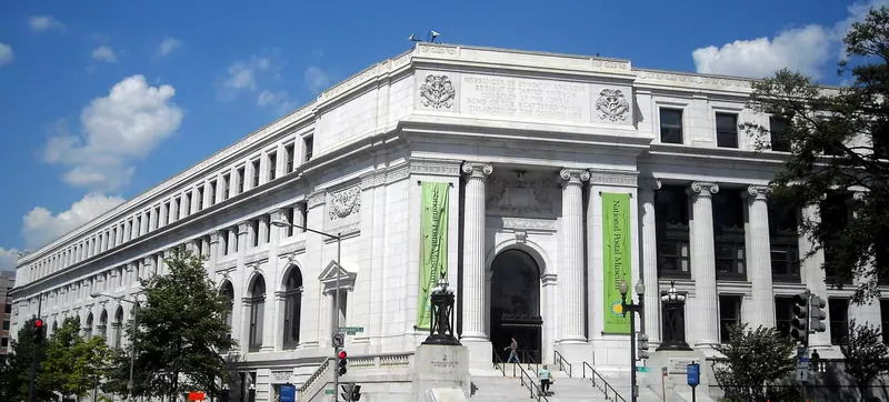 The white stone Postal Square Building in Washington, D.C. under a blue sky, with tall columns, an arched entrance, green National Postal Museum banners and a few people on the steps