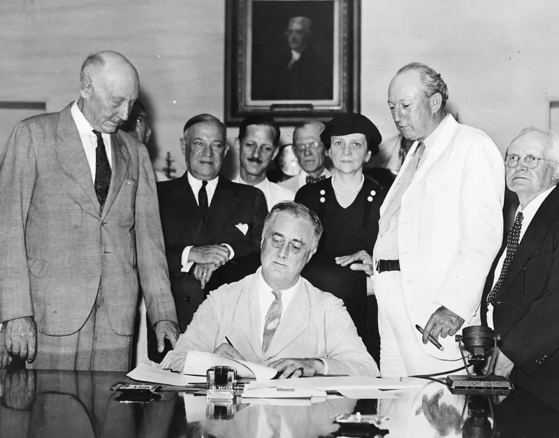Black and white photo of President Franklin D. Roosevelt seated at a desk signing a document, with lawmakers and Frances Perkins standing behind him
