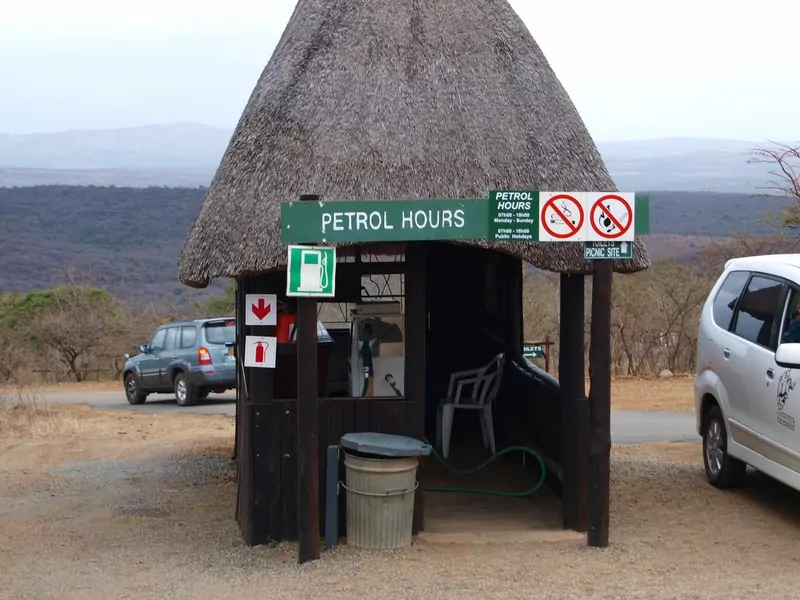 A small petrol stop under a thatched roof with a green Petrol Hours sign and no-smoking signs, with bush country and hills behind