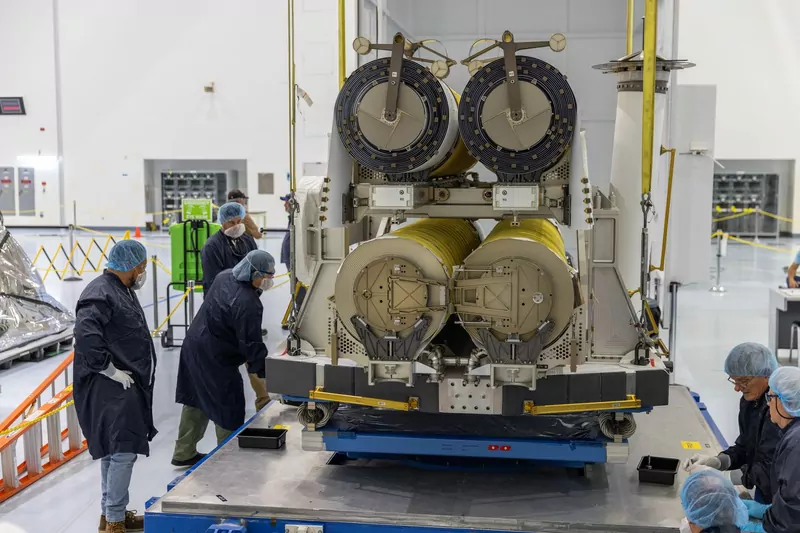 Technicians in blue lab coats and hair nets guide a crane lowering a pair of roll-out solar arrays, wound into tight rolls on a gray frame, onto a blue platform inside a bright white processing hall