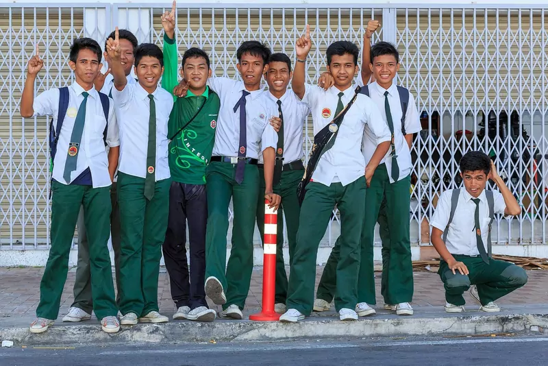 Nine Malaysian schoolboys in white short-sleeved shirts, ties and green trousers pose together on a pavement, most smiling and some pointing a finger upward, in front of a shuttered shop