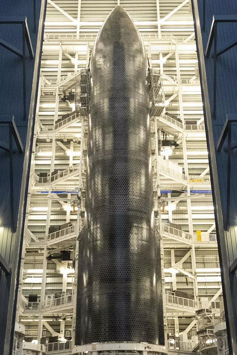 A tall Starship upper stage covered in black hexagonal heat-shield tiles, standing inside a lit hangar