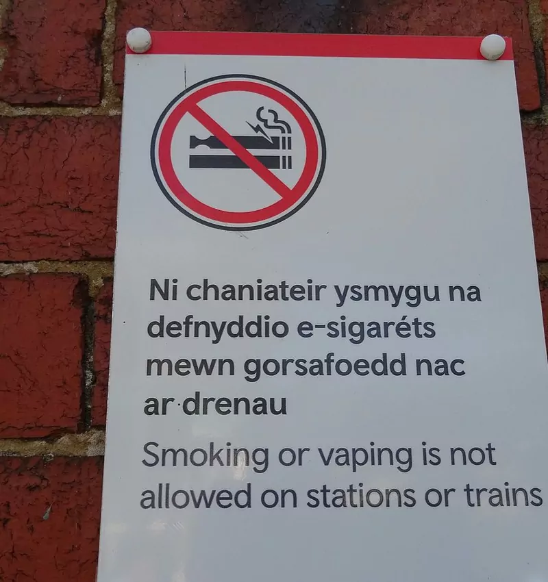 A white bilingual Welsh and English sign on a brick wall with a no-smoking symbol, reading Smoking or vaping is not allowed on stations or trains