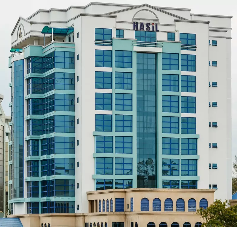 The tall white and teal glass Wisma Hasil building of the Inland Revenue Board in Kota Kinabalu, with HASiL in blue letters near the top and a beige base with arched windows