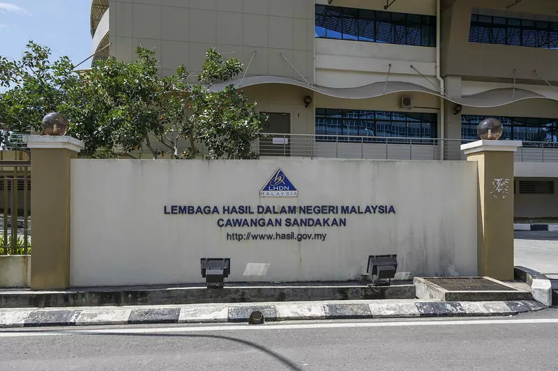 A cream wall at the entrance of a Lembaga Hasil Dalam Negeri Malaysia (LHDN) branch in Sandakan, with the blue LHDN triangle logo and the office name in dark blue letters, a tree and a beige building behind