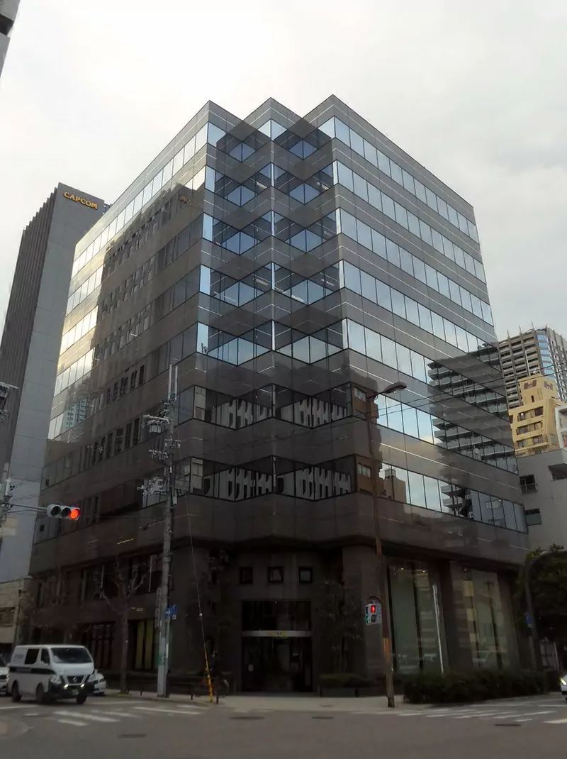 A tall dark glass office building on a street corner in Osaka, with a taller building carrying the Capcom sign behind it
