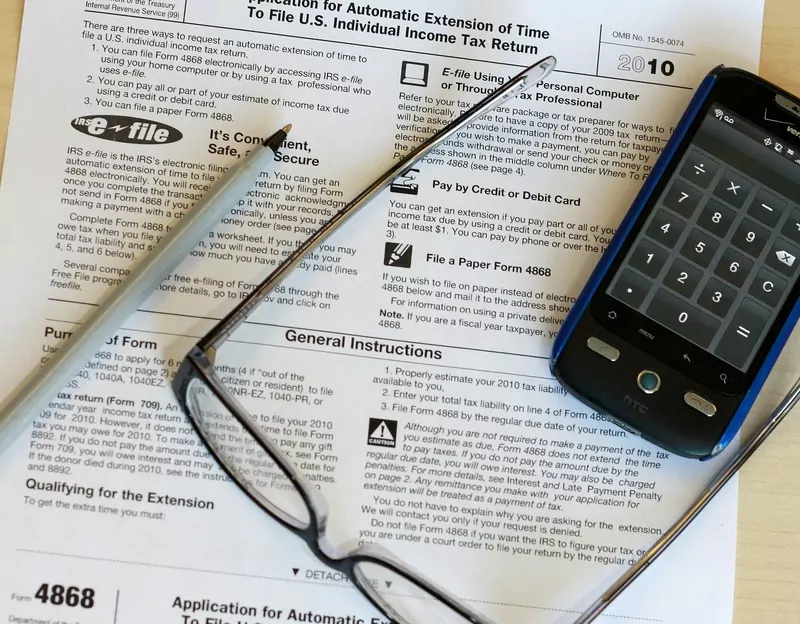 Part of IRS Form 4868 lying on a table with a pair of reading glasses and a phone showing a calculator
