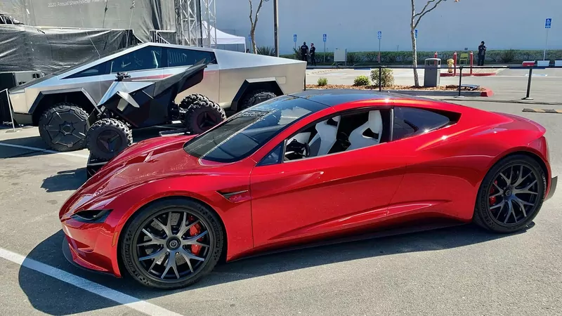 A red Tesla Roadster prototype parked beside a silver Cybertruck and a black Cyberquad on an asphalt lot, with bare trees and a white wall behind