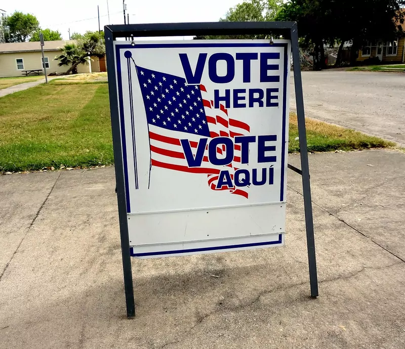 A folding metal sign on a sidewalk with an American flag and the words Vote Here and Vote Aquí