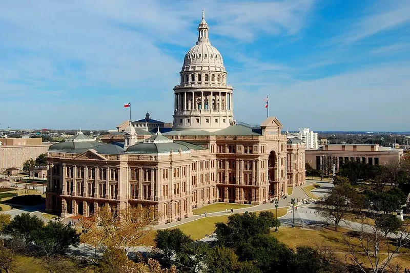 The pink granite Texas State Capitol with its tall dome, surrounded by lawns and trees under a blue sky