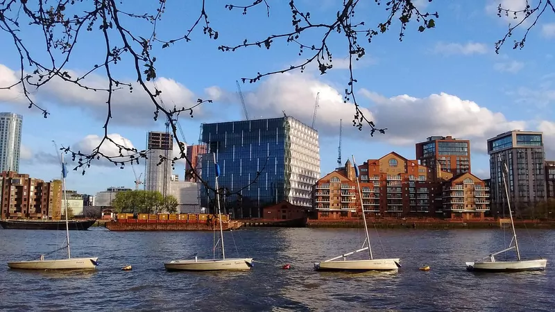 A tall glass cube building with plastic sails beside the River Thames at dusk, with cranes behind it