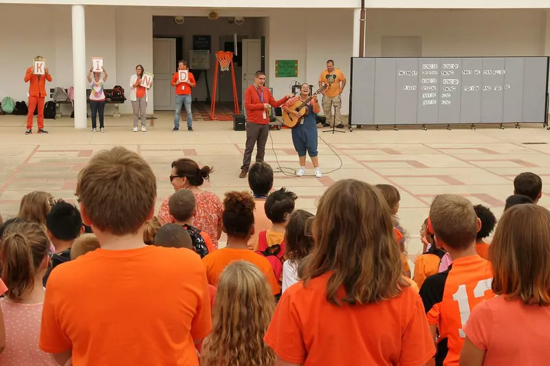 Children in orange watch a music teacher with a guitar and staff holding up letters that spell KIND in a school courtyard
