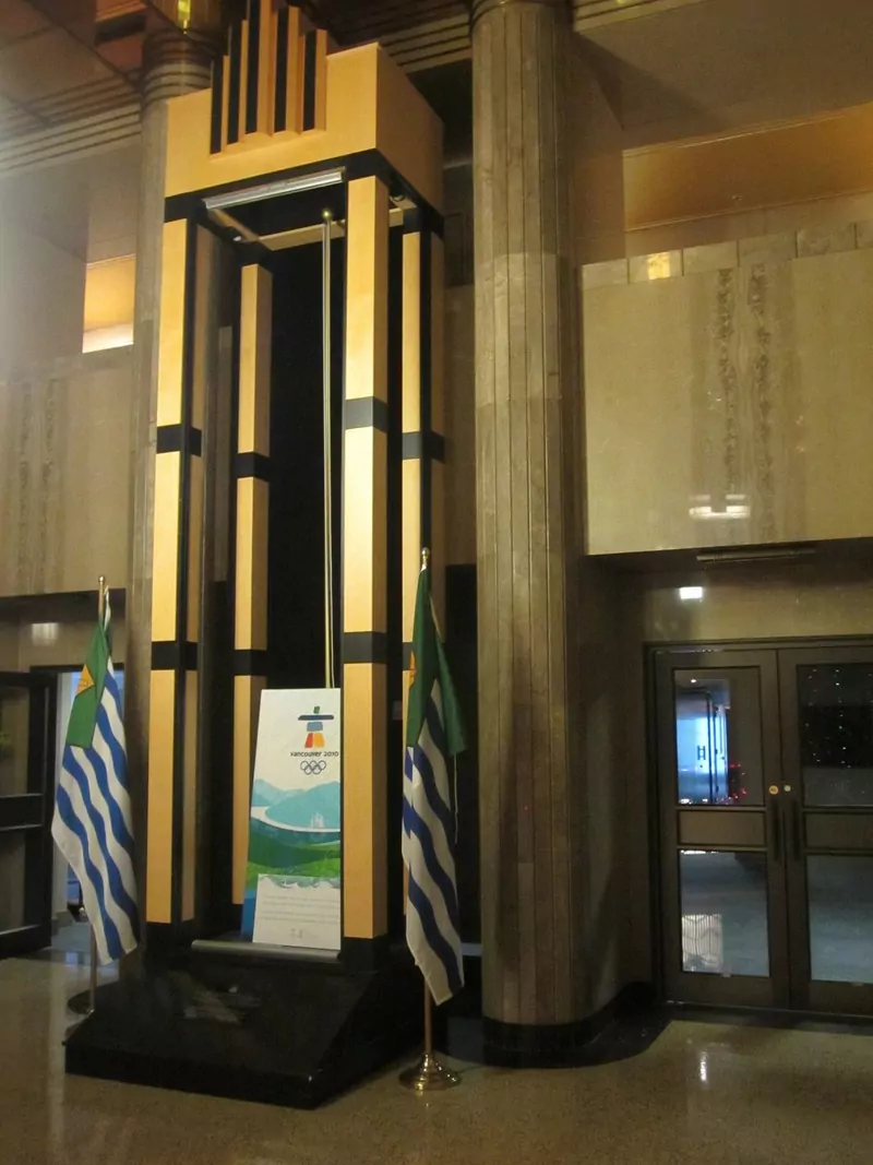 The lobby of Vancouver City Hall with a tall gold and black art deco column, blue and white Vancouver flags and polished stone floors