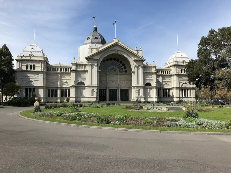 The cream Royal Exhibition Building in Melbourne with its central dome and arched entrance, behind a lawn and garden beds